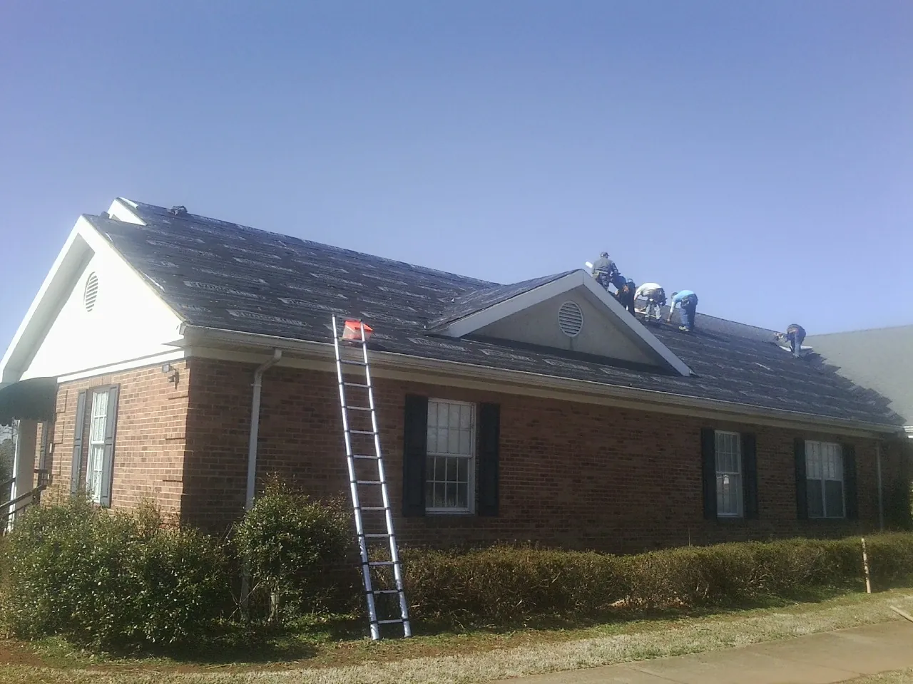 Crew tearing shingles off a brick ranch roof with a ladder set — Emergency Roof Repair work by our crew in Chappell Hill