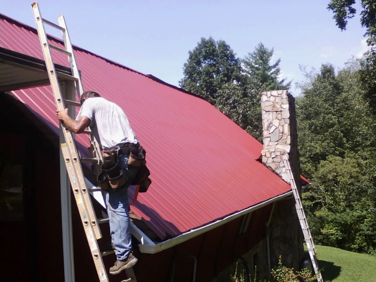 Roofer on a ladder beside a red metal roof and stone chimney — Soffit & Fascia Repair by Roofing Brothers in Chappell Hill